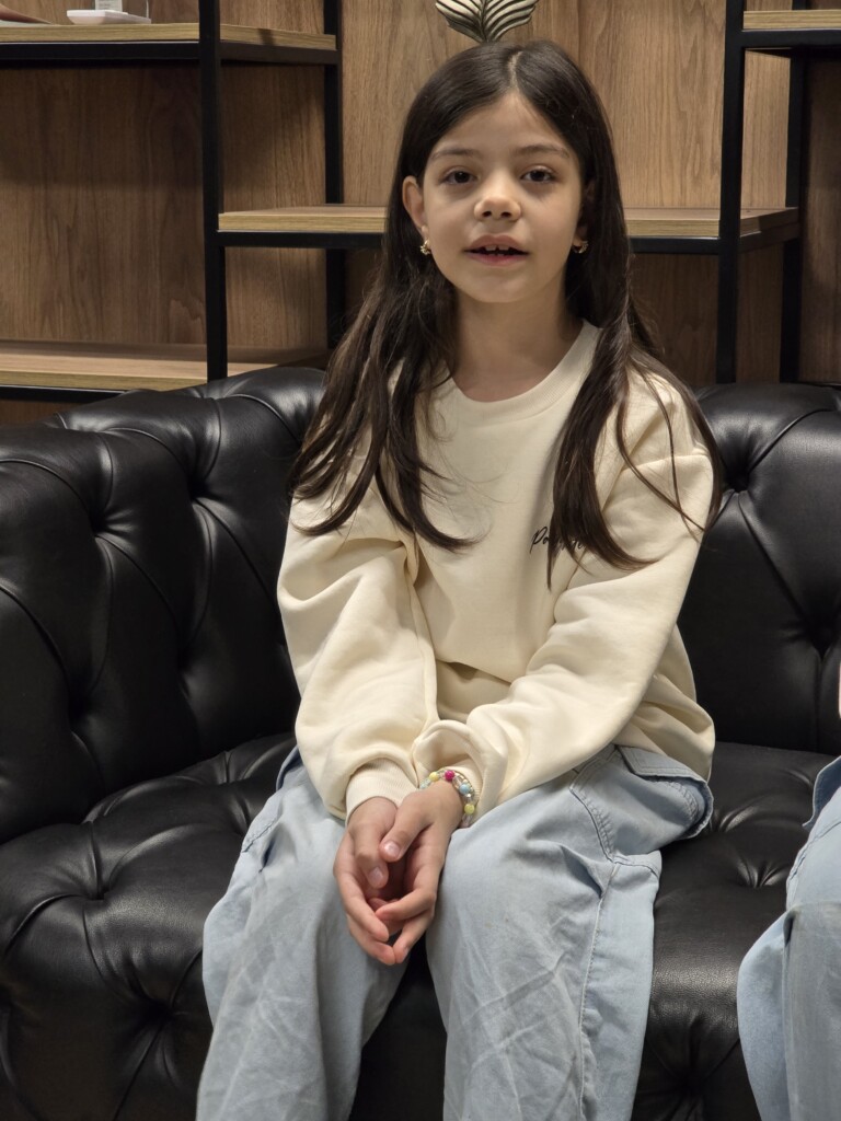 A young girl with long hair wearing a cream-colored sweatshirt is sitting on a black leather couch, looking at the camera.