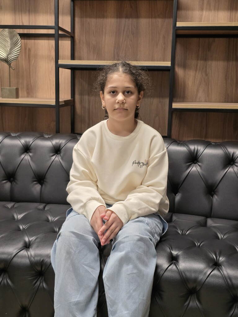 A child sitting on a black tufted couch in front of a wooden bookshelf, looking directly at the camera.