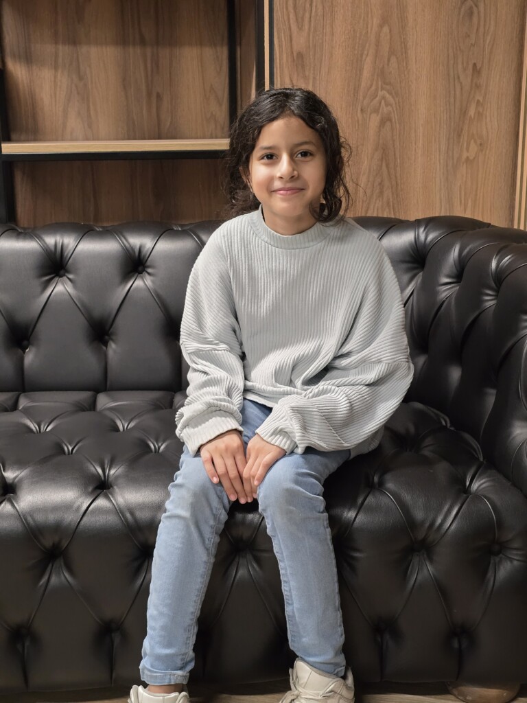 A young girl with curly hair wearing a light gray oversized sweater and jeans, sitting on a black tufted couch and smiling at the camera.