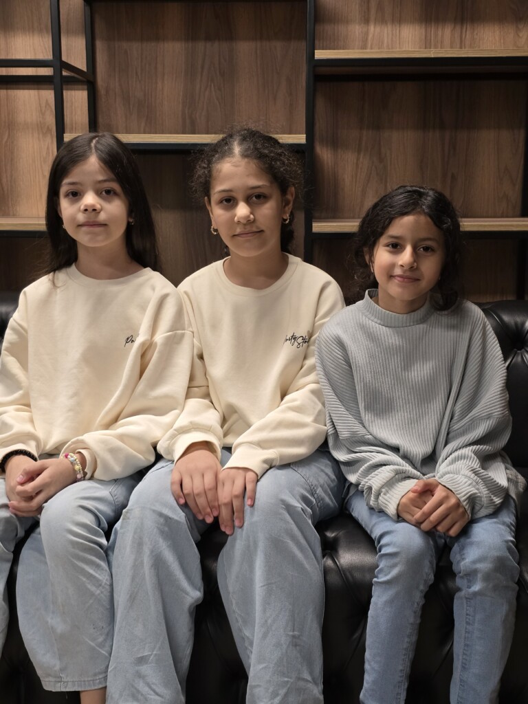 Three young girls sitting together on a black couch in a cozy setting, wearing casual sweaters and denim jeans.
