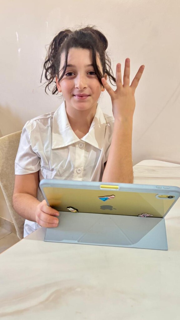 A young girl in a white shirt smiles and holds up four fingers while sitting at a table with a tablet in front of her.